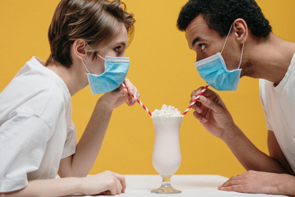 Two people wearing face masks trying to drink a milkshake through straws with a yellow background