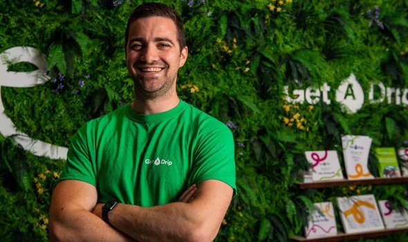 Rick, the owner, wearing a green top with the get a drip logo on standing in front of a get a drip branded wall